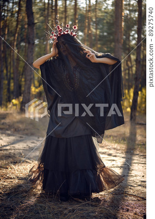 Witch in black, long dress, with red crown in her long hair under a black veil. Posing in pine forest. Spells, magic and witchcraft. Full length. 89275740