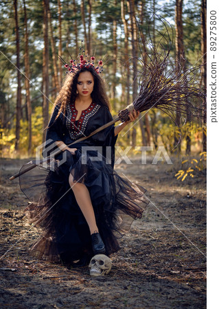 Witch in black, long dress, with red crown in her long hair. Posing with broom and skull in pine forest. Spells, magic and witchcraft. Full length. 89275800