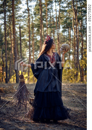 Witch in black, long dress, with red crown in her long hair. Posing with broom and skull in pine forest. Spells, magic and witchcraft. Full length. Witch in black, long dress, with red crown in her long hair. Posing with broom and skull in pine forest. Spells, magic and witchcraft. Full length. 89275802