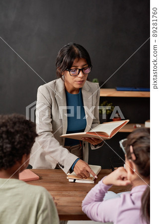 Serious young Mixed race teacher holding open textbook and explaining new topic to kids in classroom 89281760