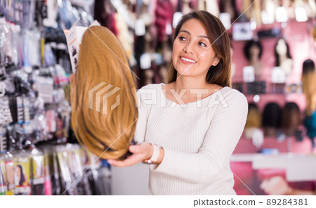 Positive young woman choosing natural wig at specialty boutique Positive young woman choosing natural wig at specialty boutique 89284381