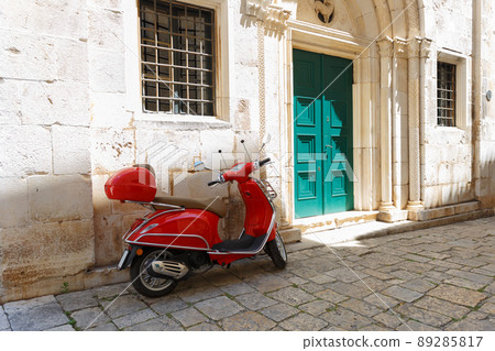 Red moped (scooter) is parked near the entrance to the building. Croatia, city of Trogir, sunny summer day. Paved road, beige walls. Selective focus, close-up. 89285817