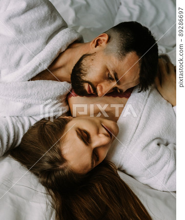 Portrait of young loving couple in bathrobes relaxing on bed at hotel 89286697