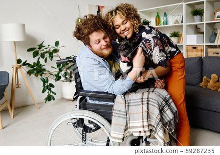 Young bearded man in wheelchair spending rehabilitation at home together with his wife and lovely dog Young bearded man in wheelchair spending rehabilitation at home together with his wife and lovely dog 89287235