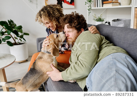 Cute dog licking face of owner while he lying on sofa with girl laughing in background Cute dog licking face of owner while he lying on sofa with girl laughing in background 89287331