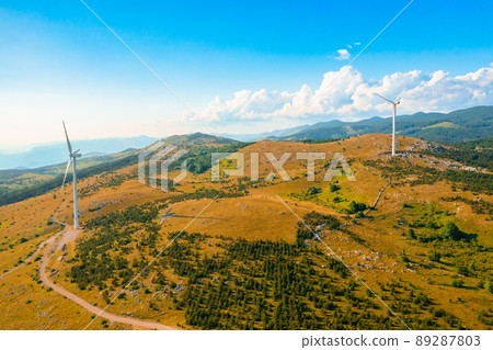Wind turbines generate alternative clean energy standing on hills tops with bushes in highland against forestry mountains on sunny day aerial view 89287803