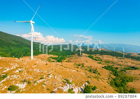 Windmills with large propellers produce green energy in highland against forestry mountains under blue sky on sunny summer day aerial view Windmills with large propellers produce green energy in highland against forestry mountains under blue sky on sunny summer day aerial view 89287804