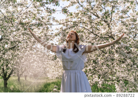 Fantasy woman in long white elegant fashion long dress walks in green spring blossom cherry garden. Happy cheerful girl princess bride. Skirt fabric flies flowing waving in wind motion. 89288722