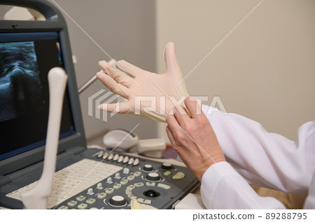 Closeup hands of radiologist, putting on sterile surgical gloves before medical examination on modern ultrasound diagnostic machine in wellness clinic Closeup hands of radiologist, putting on sterile surgical gloves before medical examination on modern ultrasound diagnostic machine in wellness clinic 89288795