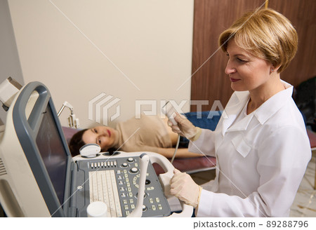 Mature female radiologist holds an ultrasound probe in hand and looks at the monitor screen while examining a patient on a modern ultrasound machine. Mature female radiologist holds an ultrasound probe in hand and looks at the monitor screen while examining a patient on a modern ultrasound machine. 89288796