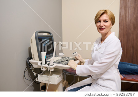 Pleasant adult European woman, radiologist, diagnostics specialist, sitting at ultrasound machine, holding transducer, looking confidently at camera Pleasant adult European woman, radiologist, diagnostics specialist, sitting at ultrasound machine, holding transducer, looking confidently at camera 89288797
