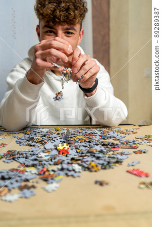 Caucasian young man with curly hair dressed in a white sweatshirt assembling a puzzle in the living room at home. training concept. Caucasian young man with curly hair dressed in a white sweatshirt assembling a puzzle in the living room at home. training concept. 89289387