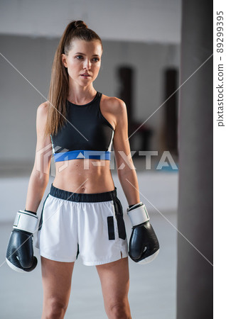 Portrait of a young attractive girl boxer in the gym wearing gloves next to a punching bag 89299395