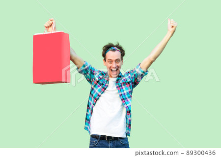Satisfied modern young shopoholic man in white t-shirt and checkered shirt triumping with raised hands up with shopping bags and opened mouth, looking at camera. Studio shot Satisfied modern young shopoholic man in white t-shirt and checkered shirt triumping with raised hands up with shopping bags and opened mouth, looking at camera. Studio shot 89300436