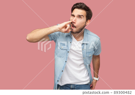 Portrait of crazy handsome bearded young man in blue casual style shirt standing and drilling his nose with funny face. indoor studio shot, isolated on pink background. Portrait of crazy handsome bearded young man in blue casual style shirt standing and drilling his nose with funny face. indoor studio shot, isolated on pink background. 89300482