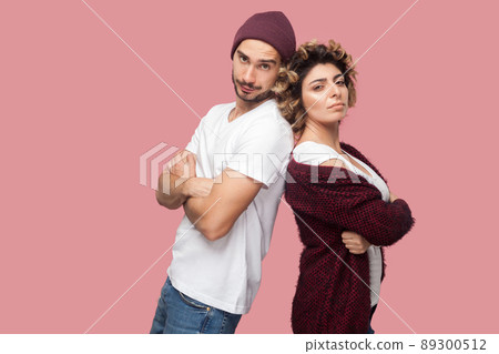 Portrait of serious couple of friends in casual style standing back to back, crossing arms and looking at camera with proud face. Isolated, indoor, studio shot, pink background 89300512