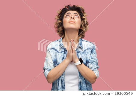 My god please help me. Portrait of hopeful young woman with curly hairstyle in casual blue shirt standing with palm hand and praying or begging. indoor studio shot, isolated on pink background. My god please help me. Portrait of hopeful young woman with curly hairstyle in casual blue shirt standing with palm hand and praying or begging. indoor studio shot, isolated on pink background. 89300549
