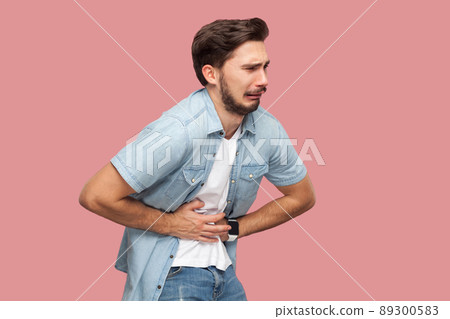 Stomach pain. Portrait of sick sad handsome bearded young man in blue casual style shirt standing and holding his painful belly. indoor studio shot, isolated on pink background. 89300583