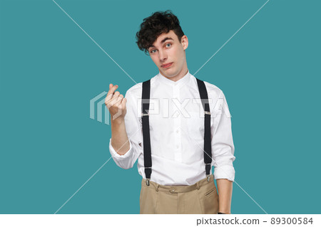 Worry or hopeful man showing money gesture. Portrait of handsome hipster curly young businessman in classic casual white shirt and suspender standing. indoor studio shot isolated on blue background. 89300584