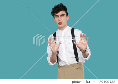 shocked or scared man blocking with hands and looking at camera. Portrait of handsome hipster curly young businessman in classic casual white shirt, suspender. studio shot isolated on blue background. shocked or scared man blocking with hands and looking at camera. Portrait of handsome hipster curly young businessman in classic casual white shirt, suspender. studio shot isolated on blue background. 89300588