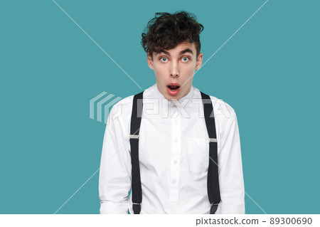 Shocked man looking at camera with big eyes and open mouth. Portrait of handsome hipster curly young businessman in classic casual white shirt and suspender. studio shot isolated on blue background. 89300690