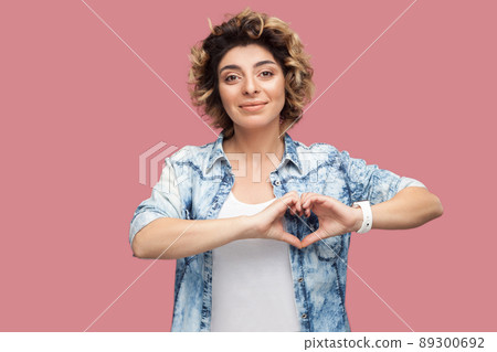 Portrait of happy young woman with curly hairstyle in casual blue shirt standing with heart hands shape gesture and looking at camera smiling. indoor studio shot, isolated on pink background. 89300692