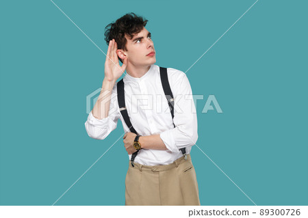 Attentive man trying to hear with hand near ears. Portrait of handsome hipster curly young businessman in classic casual white shirt, suspender standing. indoor studio shot isolated on blue background 89300726