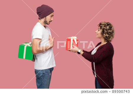 Side view portrait of amazing couple of friends in casual style standing, girl giving present box, boy hide behind gift, celebrate anniversary. Isolated, indoor, studio shot, pink background Side view portrait of amazing couple of friends in casual style standing, girl giving present box, boy hide behind gift, celebrate anniversary. Isolated, indoor, studio shot, pink background 89300743