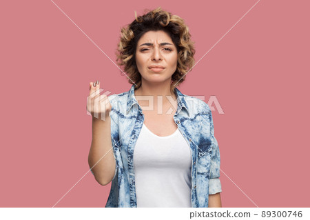 Portrait of serious young woman with curly hairstyle in casual blue shirt standing with money or italian gesture and looking at camera. indoor studio shot, isolated on pink background. 89300746