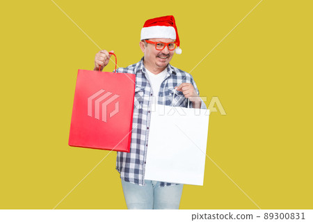 Happy modern middle aged man in casual style and red new year santa cap, eyeglasses and shirt, holding shopping packages with toothy smile, looking at camera. indoor, isolated on yellow background 89300831