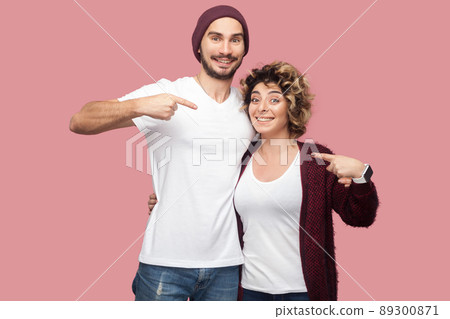 Funny couple of friends in casual style standing, hugging and pointing fingers to each other, looking at camera with happiness and toothy smile. Isolated, indoor, studio shot, pink background Funny couple of friends in casual style standing, hugging and pointing fingers to each other, looking at camera with happiness and toothy smile. Isolated, indoor, studio shot, pink background 89300871