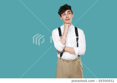 Dreamy man standing, dreaming and looking away. Portrait of handsome hipster curly young businessman in classic casual white shirt, suspender standing. indoor studio shot isolated on blue background. Dreamy man standing, dreaming and looking away. Portrait of handsome hipster curly young businessman in classic casual white shirt, suspender standing. indoor studio shot isolated on blue background. 89300932