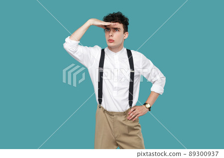 Attentive man looking to far with hand on forrhead. Portrait of handsome hipster curly young businessman in classic casual white shirt and suspender standing. studio shot isolated on blue background. Attentive man looking to far with hand on forrhead. Portrait of handsome hipster curly young businessman in classic casual white shirt and suspender standing. studio shot isolated on blue background. 89300937