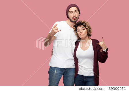 Portrait of funny couple of friends in casual style standing, hugging each other and showing rock and roll hand sign gesture, looking at camera. Isolated, indoor, studio shot, pink background 89300951