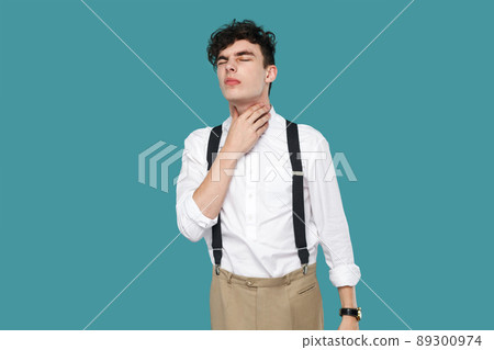 Throat pain. man holding his painful neck and feeling bad. Portrait of handsome hipster young businessman in classic casual white shirt and suspender. indoor studio shot isolated on blue background. 89300974