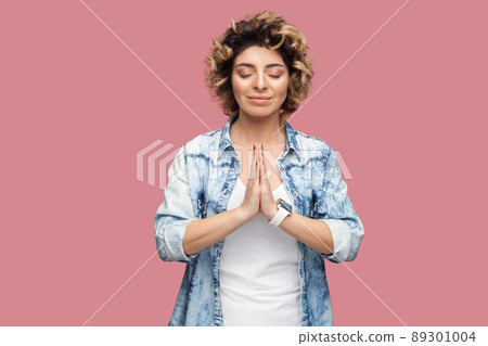 Portrait of calm young woman with curly hairstyle in casual blue shirt standing with closed eyes, palm hands, smile and doing yoga meditating. indoor studio shot, isolated on pink background. Portrait of calm young woman with curly hairstyle in casual blue shirt standing with closed eyes, palm hands, smile and doing yoga meditating. indoor studio shot, isolated on pink background. 89301004