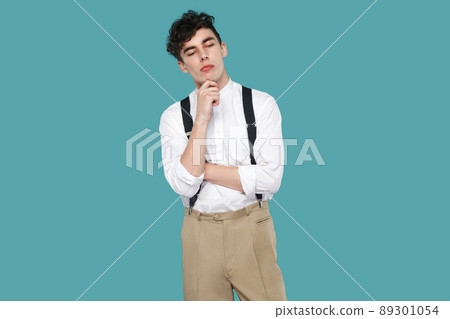 thoughtful man, touching his chin and thinking what to do. Portrait of handsome hipster curly young businessman in classic casual white shirt and suspender. studio shot isolated on blue background. thoughtful man, touching his chin and thinking what to do. Portrait of handsome hipster curly young businessman in classic casual white shirt and suspender. studio shot isolated on blue background. 89301054