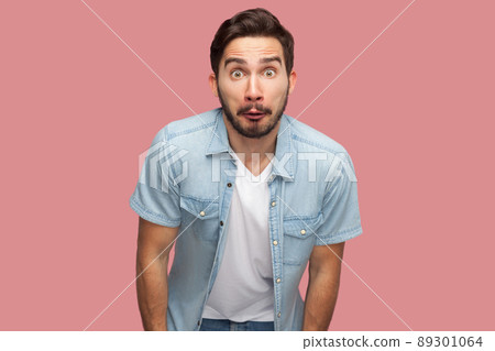 Portrait of surprised handsome bearded young man in blue casual style shirt standing and looking at camera with shocked unbelievable face. indoor studio shot, isolated on pink background. 89301064