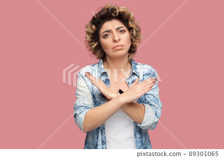 Portrait of worry young woman with curly hairstyle in casual blue shirt standing with X sign or end gesture, and looking at camera with sad face. indoor studio shot, isolated on pink background. 89301065