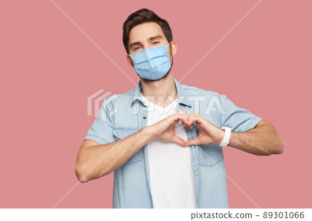 Portrait of happy smiley young man with surgical medical mask in blue shirt standing with love hand gesture on heart and looking at camera smiling. indoor studio shot, isolated on pink background. 89301066
