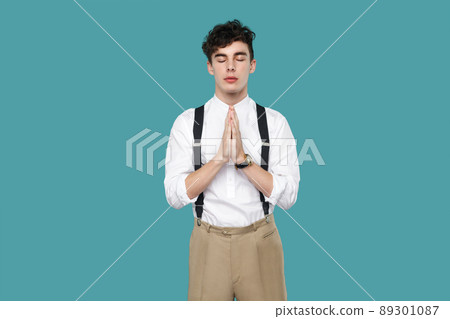 Calm relaxed man closed eyes in yoga namaste pose. Portrait of handsome hipster curly young businessman in classic casual white shirt and suspender standing. studio shot isolated on blue background. Calm relaxed man closed eyes in yoga namaste pose. Portrait of handsome hipster curly young businessman in classic casual white shirt and suspender standing. studio shot isolated on blue background. 89301087