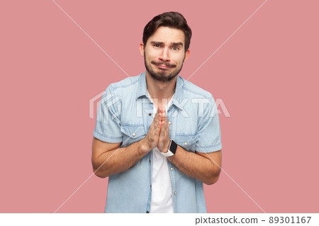 Please help me, or forgive. Portrait of sad hopeful handsome bearded young man in blue casual style shirt standing, looking at camera and begging. indoor studio shot, isolated on pink background. 89301167