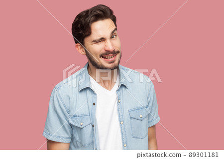 Portrait of funny handsome bearded young man in blue casual style shirt standing, winking and looking at camera with funny face and toothy smile. indoor studio shot, isolated on pink background. 89301181