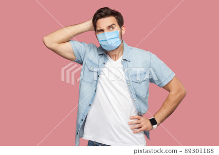 Portrait of young man with surgical medical mask in blue casual style shirt standing, scratching his head, looking away and thinking what to do. indoor studio shot, isolated on pink background. 89301188