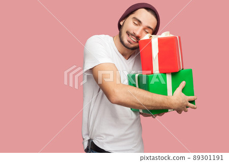 Portrait of happy satisfied handsome bearded young hipster man in white shirt and casual hat standing and holding two present bo and hugging it. Indoor, isolated, studio shot, pink background 89301191