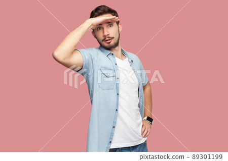 Portrait of attentive handsome bearded young man in blue casual style shirt standing with hand on forehead and looking away. indoor studio shot, isolated on pink background. Portrait of attentive handsome bearded young man in blue casual style shirt standing with hand on forehead and looking away. indoor studio shot, isolated on pink background. 89301199