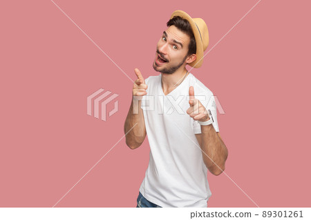 Portrait of surprised bearded young man in white shirt with hat standing, pointing and looking at camera with amazed face. indoor studio shot, isolated on pink background copyspace. Portrait of surprised bearded young man in white shirt with hat standing, pointing and looking at camera with amazed face. indoor studio shot, isolated on pink background copyspace. 89301261