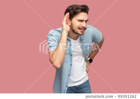 Portrait of attentive handsome bearded young man in blue casual style shirt standing with hand on ear and trying to hear something with serious face. indoor studio shot, isolated on pink background. 89301262
