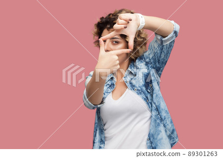 Portrait of serious attentive young woman with curly hairstyle in casual blue shirt standing with crop hands composition and looking at her target. indoor studio shot, isolated on pink background. Portrait of serious attentive young woman with curly hairstyle in casual blue shirt standing with crop hands composition and looking at her target. indoor studio shot, isolated on pink background. 89301263