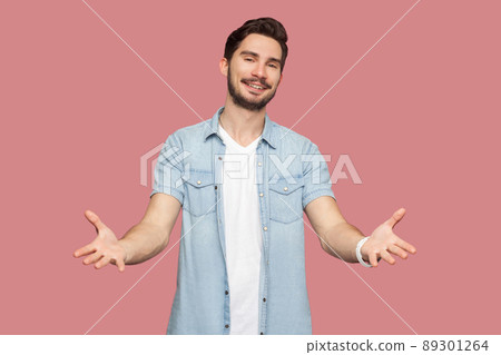 Portrait of happy handsome bearded young man in blue casual style shirt standing with raised arms and looking at camera with toothy smile. indoor studio shot, isolated on pink background. 89301264